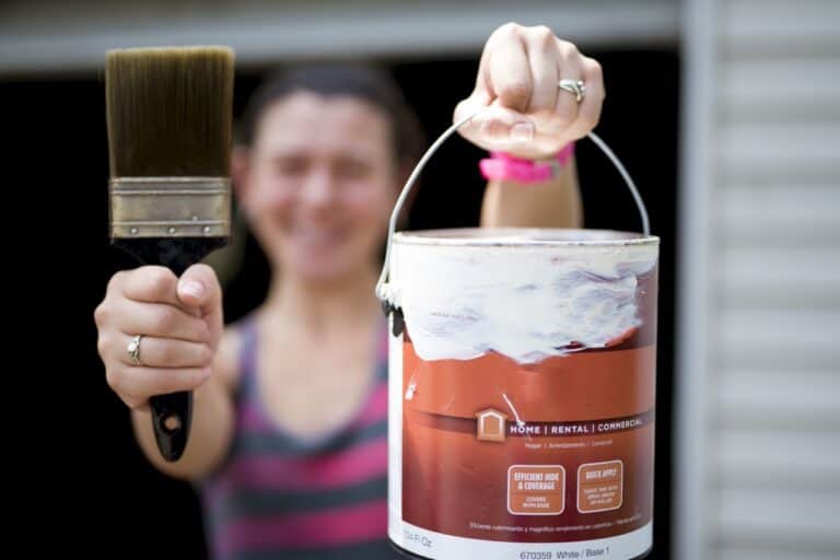 A closeup shot of a female holding up a paint can and a paintbrush with a blurred background