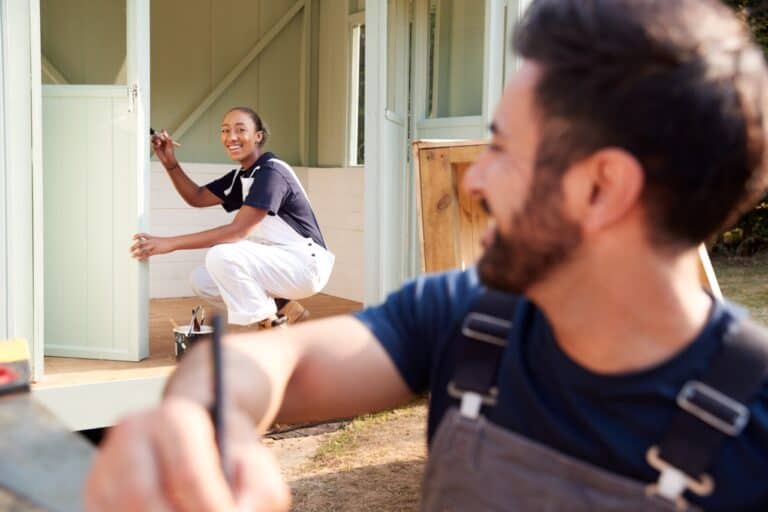 Male Carpenter With Female Apprentice Measuring Wood To Build Outdoor Summerhouse In Garden